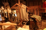 A vintage sewing room features a wooden mannequin wearing a tailored vest. Several sewing machines are placed on wooden tables, surrounded by fabric and paper patterns. The space has an industrial yet cozy ambiance, accentuated by warm lighting and wrought iron railings.