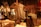 A vintage sewing room features a wooden mannequin wearing a tailored vest. Several sewing machines are placed on wooden tables, surrounded by fabric and paper patterns. The space has an industrial yet cozy ambiance, accentuated by warm lighting and wrought iron railings.