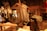 A vintage sewing room features a wooden mannequin wearing a tailored vest. Several sewing machines are placed on wooden tables, surrounded by fabric and paper patterns. The space has an industrial yet cozy ambiance, accentuated by warm lighting and wrought iron railings.