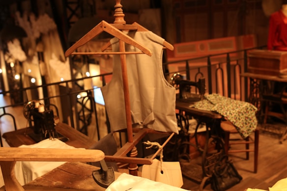 A vintage sewing room features a wooden mannequin wearing a tailored vest. Several sewing machines are placed on wooden tables, surrounded by fabric and paper patterns. The space has an industrial yet cozy ambiance, accentuated by warm lighting and wrought iron railings.