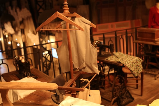 A vintage sewing room features a wooden mannequin wearing a tailored vest. Several sewing machines are placed on wooden tables, surrounded by fabric and paper patterns. The space has an industrial yet cozy ambiance, accentuated by warm lighting and wrought iron railings.