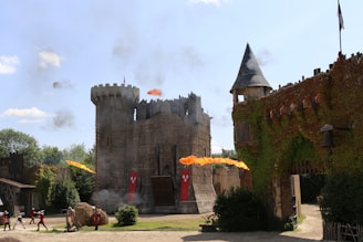 A medieval-style castle surrounded by greenery, with flames and smoke indicating a staged battle. Archers with bows, soldiers, and the architecture display a historical reenactment setting.
