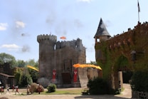 A medieval-style castle surrounded by greenery, with flames and smoke indicating a staged battle. Archers with bows, soldiers, and the architecture display a historical reenactment setting.