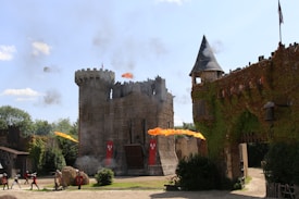 A medieval-style castle surrounded by greenery, with flames and smoke indicating a staged battle. Archers with bows, soldiers, and the architecture display a historical reenactment setting.