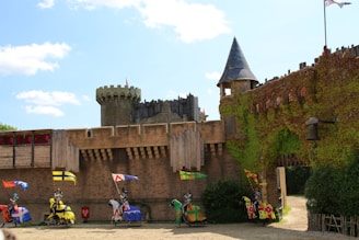 Five armored knights on horseback, each carrying colorful medieval banners, participate in a jousting tournament in front of an ancient stone castle. The castle features ivy-covered walls, a turret, and wooden battlements.