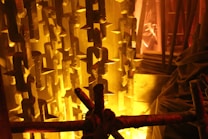 Rows of heavy, industrial chains hang vertically against a backdrop illuminated by warm, yellowish light. The texture of the chains is rugged, and they create a dramatic effect with sharp, pointed shapes. Draped fabric and structural elements are present, adding layers and depth to the scene.