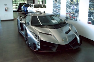 A sleek, silver luxury sports car is prominently displayed in an indoor showroom. The car has a futuristic and aerodynamic design with sharp angles and glossy finishes. Behind it, two more vehicles in blue and white are partially visible. The walls are adorned with automotive-themed artwork, and there's ample natural light filtering through large windows.