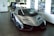 A sleek, silver luxury sports car is prominently displayed in an indoor showroom. The car has a futuristic and aerodynamic design with sharp angles and glossy finishes. Behind it, two more vehicles in blue and white are partially visible. The walls are adorned with automotive-themed artwork, and there's ample natural light filtering through large windows.