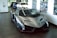 A sleek, silver luxury sports car is prominently displayed in an indoor showroom. The car has a futuristic and aerodynamic design with sharp angles and glossy finishes. Behind it, two more vehicles in blue and white are partially visible. The walls are adorned with automotive-themed artwork, and there's ample natural light filtering through large windows.