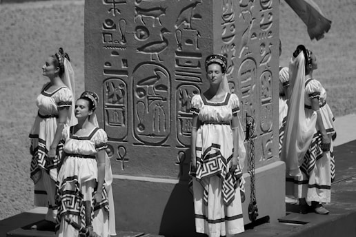 Women dressed in ancient-style garments stand near an obelisk adorned with hieroglyphs. The garments have a Greek or Egyptian influence, featuring headbands and veils. The background suggests a historical or cultural setting, possibly outdoors.
