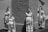 Women dressed in ancient-style garments stand near an obelisk adorned with hieroglyphs. The garments have a Greek or Egyptian influence, featuring headbands and veils. The background suggests a historical or cultural setting, possibly outdoors.