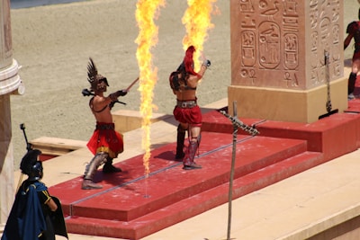 Two people wearing gladiator-style costumes perform near a flaming pole in an ancient-themed setting. One holds a sword while the other is mid-action, both adorned in red attire with helmets. In the background, hieroglyphs are visible on a large pillar.