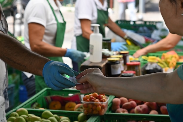 People are engaged in a transaction at a market stall. The scene includes fruits and vegetables, bottles of preserves, and a bottle of hand sanitizer on the counter. Two individuals are in focus, exchanging money or a receipt, while others can be seen working in the background wearing gloves and aprons.
