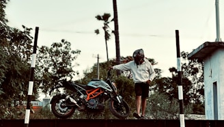 A person wearing a helmet and a casual outfit stands next to a sports motorcycle on a platform. The background features lush greenery and tall trees, creating a natural setting. There are black and white striped poles on either side, and a building structure is visible on the right.