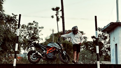 A person wearing a helmet and a casual outfit stands next to a sports motorcycle on a platform. The background features lush greenery and tall trees, creating a natural setting. There are black and white striped poles on either side, and a building structure is visible on the right.