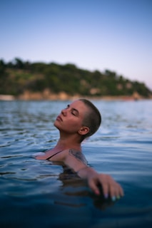 A person with short hair is floating in a body of water, with eyes closed and a serene expression. The background features a blurred view of a tree-covered hillside. The lighting suggests a natural, outdoor setting during twilight.
