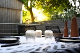 An outdoor dining table is set with clear glass tumblers, black plates, and cutlery. There are terracotta vases containing greenery and a bowl on the table. The background consists of wooden structures and lush green trees, bathed in the warm glow of sunlight.