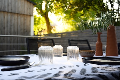 An outdoor dining table is set with clear glass tumblers, black plates, and cutlery. There are terracotta vases containing greenery and a bowl on the table. The background consists of wooden structures and lush green trees, bathed in the warm glow of sunlight.