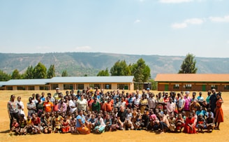 A large group of people consisting of adults and children is gathered outdoors in a rural setting. Behind them, there are several single-story buildings with colorful roofs, and a backdrop of hills and trees. The group seems to be posing for a communal photograph. The day appears to be sunny, casting bright light on the scene.