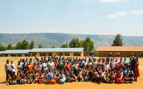 A large group of people consisting of adults and children is gathered outdoors in a rural setting. Behind them, there are several single-story buildings with colorful roofs, and a backdrop of hills and trees. The group seems to be posing for a communal photograph. The day appears to be sunny, casting bright light on the scene.