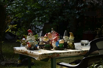 Several colorful garden gnome figurines are arranged on an outdoor table covered with a floral-patterned cloth. The scene is set in a garden with lush greenery in the background. A black plastic chair is partially visible on the right side, and a transparent ashtray can be seen on the table.