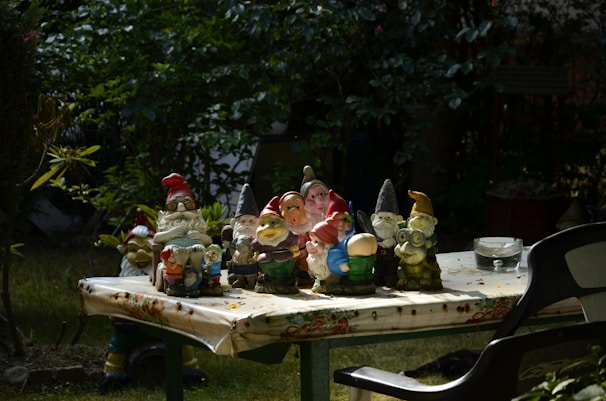 Several colorful garden gnome figurines are arranged on an outdoor table covered with a floral-patterned cloth. The scene is set in a garden with lush greenery in the background. A black plastic chair is partially visible on the right side, and a transparent ashtray can be seen on the table.