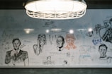 A whiteboard filled with various hand-drawn sketches and doodles, including portraits, anime characters, and what appears to be famous figures. There are abstract shapes and handwritten messages scattered across the surface. A hanging light fixture is visible above the board, illuminating the drawings.