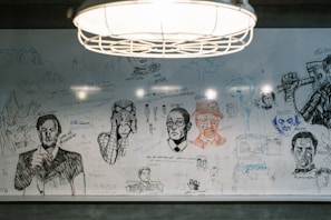 A whiteboard filled with various hand-drawn sketches and doodles, including portraits, anime characters, and what appears to be famous figures. There are abstract shapes and handwritten messages scattered across the surface. A hanging light fixture is visible above the board, illuminating the drawings.