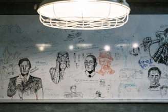 A whiteboard filled with various hand-drawn sketches and doodles, including portraits, anime characters, and what appears to be famous figures. There are abstract shapes and handwritten messages scattered across the surface. A hanging light fixture is visible above the board, illuminating the drawings.