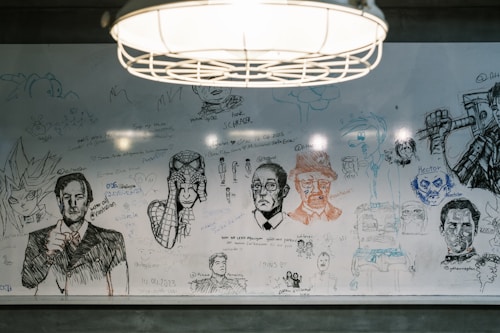A whiteboard filled with various hand-drawn sketches and doodles, including portraits, anime characters, and what appears to be famous figures. There are abstract shapes and handwritten messages scattered across the surface. A hanging light fixture is visible above the board, illuminating the drawings.