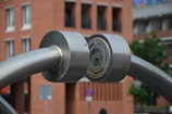 A large metallic structure with cylindrical components is prominently featured. The structure seems to be made of stainless steel, with two thick, curved rods meeting at a central circular joint. The background shows an urban setting with a brick building and blurred hints of greenery.