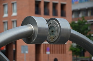 A large metallic structure with cylindrical components is prominently featured. The structure seems to be made of stainless steel, with two thick, curved rods meeting at a central circular joint. The background shows an urban setting with a brick building and blurred hints of greenery.