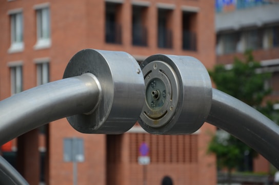 A large metallic structure with cylindrical components is prominently featured. The structure seems to be made of stainless steel, with two thick, curved rods meeting at a central circular joint. The background shows an urban setting with a brick building and blurred hints of greenery.