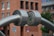 A large metallic structure with cylindrical components is prominently featured. The structure seems to be made of stainless steel, with two thick, curved rods meeting at a central circular joint. The background shows an urban setting with a brick building and blurred hints of greenery.