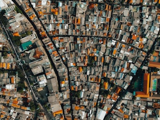 An overhead view of a densely populated urban area, featuring a grid of tightly packed buildings with varied rooftops. The structures are aligned along narrow streets, creating a maze-like pattern. A mix of rectangular and square buildings can be seen, with some larger complexes and a sports facility visible.