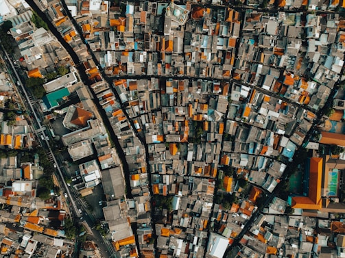 An overhead view of a densely populated urban area, featuring a grid of tightly packed buildings with varied rooftops. The structures are aligned along narrow streets, creating a maze-like pattern. A mix of rectangular and square buildings can be seen, with some larger complexes and a sports facility visible.