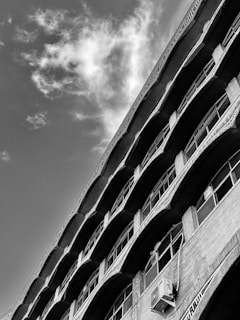 A black and white photograph of a building with a unique architectural design, featuring curved arches and multiple windows arranged in a repetitive pattern. The perspective creates a dynamic angle that emphasizes the height of the structure against a partly cloudy sky.