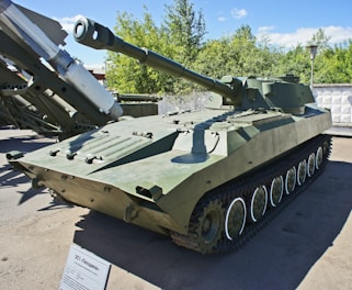 An armored military vehicle with a long barrel cannon is positioned outdoors on a paved surface. It is painted in a camouflage shade of green and features tracks for mobility. The vehicle is displayed in a museum or exhibition setting with a small informational placard in front of it. Surrounding the vehicle are some trees and a concrete wall.