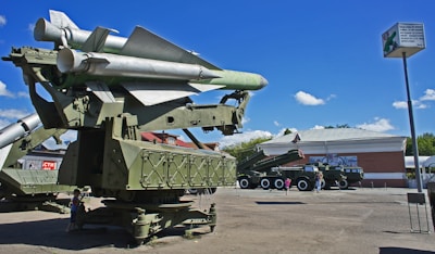 A large military missile launcher is displayed outdoors on a concrete surface. The launcher is equipped with multiple rocket tubes pointing skywards and is painted in a matte green color. In the background, a large military truck with additional equipment is visible. A few people are standing nearby, possibly visitors to a museum or a public exhibition. The setting includes a building with a large painted mural, trees, and a clear sky with fluffy clouds.