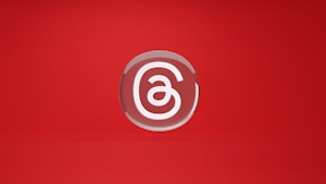 A stylized letter 'a' is encased in a circular, glossy emblem set against a vibrant red background. The design is minimalistic and centered, with a smooth, reflective surface giving it a polished appearance.