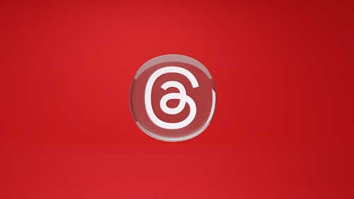 A stylized letter 'a' is encased in a circular, glossy emblem set against a vibrant red background. The design is minimalistic and centered, with a smooth, reflective surface giving it a polished appearance.