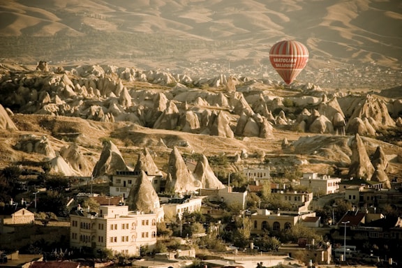 A picturesque landscape featuring unique rock formations with a combination of conical and jagged shapes. In the foreground, a small village with traditional buildings is nestled among trees. A prominent red and white striped hot air balloon is floating in the sky, providing a sense of adventure and exploration.