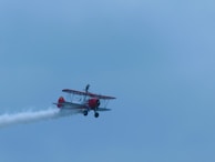 A vintage biplane with red accents flies through a clear sky, emitting a trail of white smoke. A person is standing on top of the airplane, suggesting an aerobatic airshow performance.