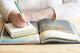 A person is writing in a small spiral notebook with a green pencil, resting on a wooden table alongside an open book displaying a scenic photograph. The individual is wearing a light-colored, striped long-sleeve shirt.