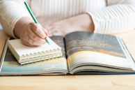 A person is writing in a small spiral notebook with a green pencil, resting on a wooden table alongside an open book displaying a scenic photograph. The individual is wearing a light-colored, striped long-sleeve shirt.