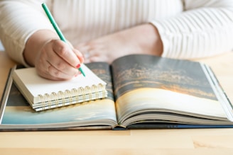 A person is writing in a small spiral notebook with a green pencil, resting on a wooden table alongside an open book displaying a scenic photograph. The individual is wearing a light-colored, striped long-sleeve shirt.