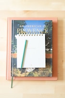 Three items, a closed book titled 'An American In Provence' by Jamie Beck at the bottom, a peach-colored closed book beneath it, and a spiral-bound notepad with a green pen on top are arranged in a neat stack on a light wooden surface.