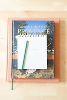Three items, a closed book titled 'An American In Provence' by Jamie Beck at the bottom, a peach-colored closed book beneath it, and a spiral-bound notepad with a green pen on top are arranged in a neat stack on a light wooden surface.