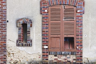 A weathered wall with two different windows. On the left, a narrow, arched window is framed with multicolored bricks and partially filled with clear glass blocks. On the right, a larger wooden shuttered window is seen, with broken sections revealing a dark interior behind it. The wall has visible areas of peeling plaster and rough textures.