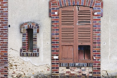 A weathered wall with two different windows. On the left, a narrow, arched window is framed with multicolored bricks and partially filled with clear glass blocks. On the right, a larger wooden shuttered window is seen, with broken sections revealing a dark interior behind it. The wall has visible areas of peeling plaster and rough textures.